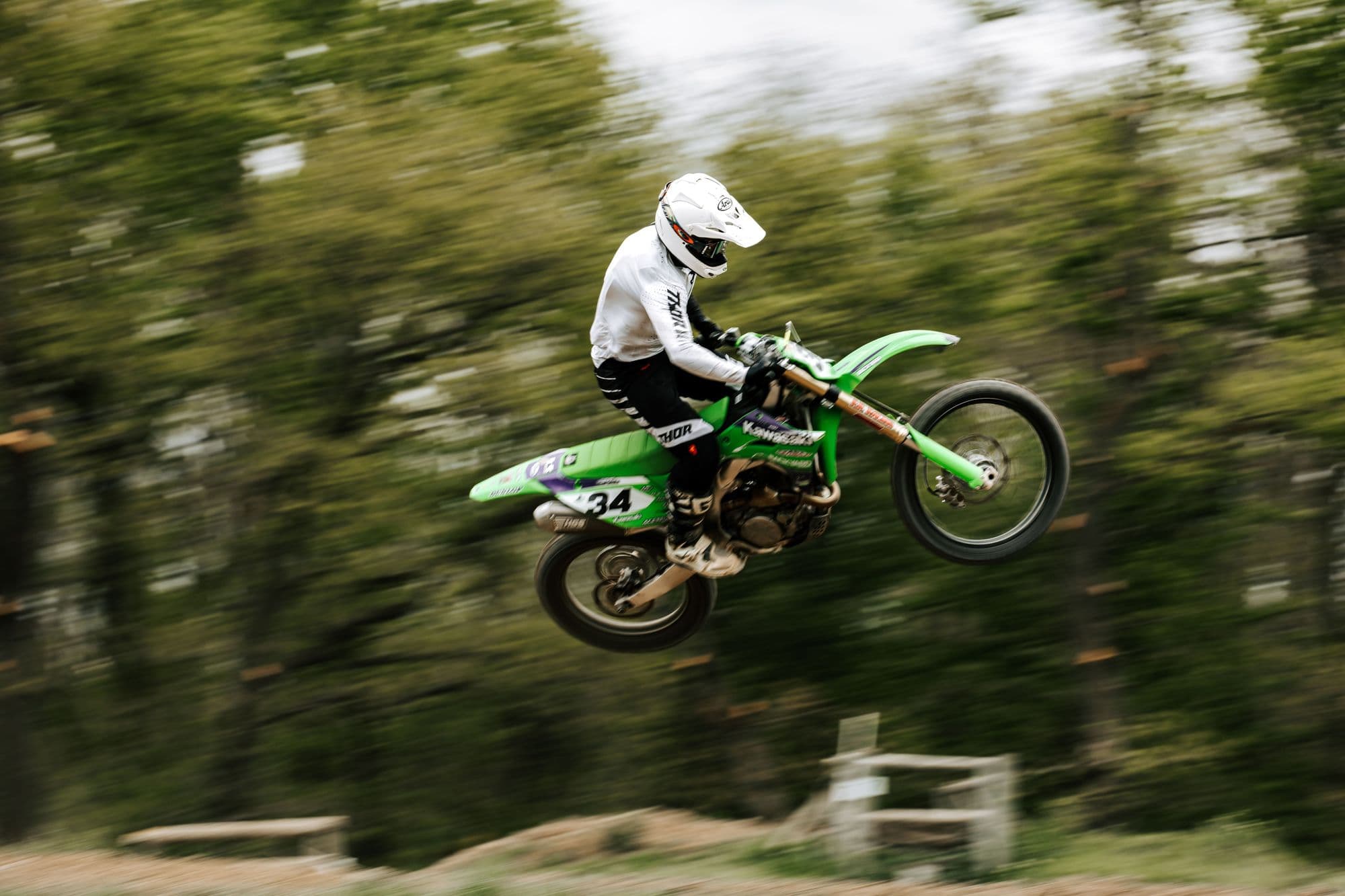 Rider on a green bike airborne over a jump, trees blurred behind