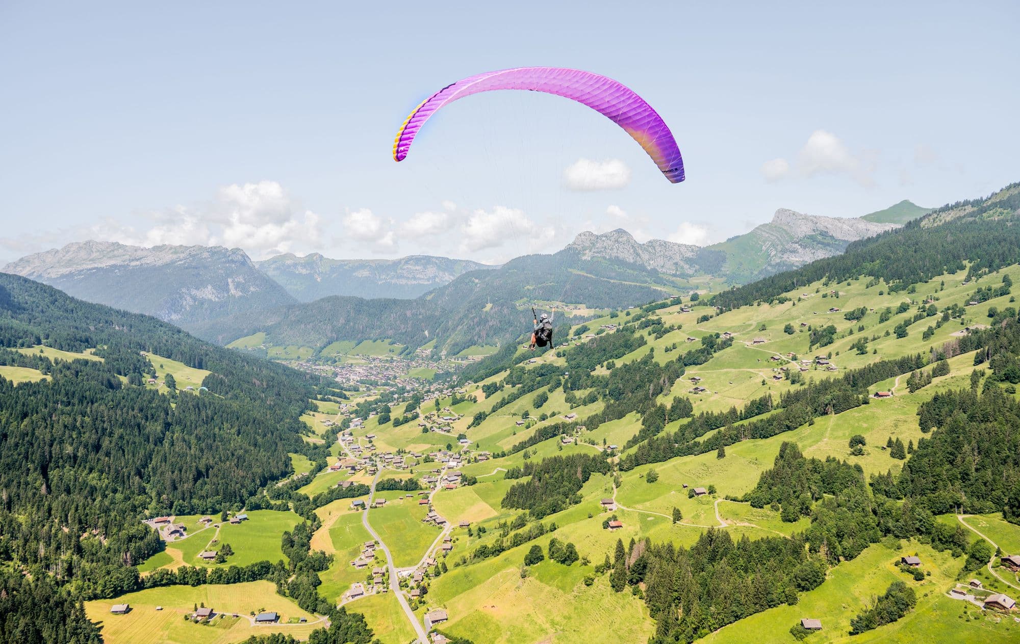 A paraglider under a purple wing flying high above a green alpine valley