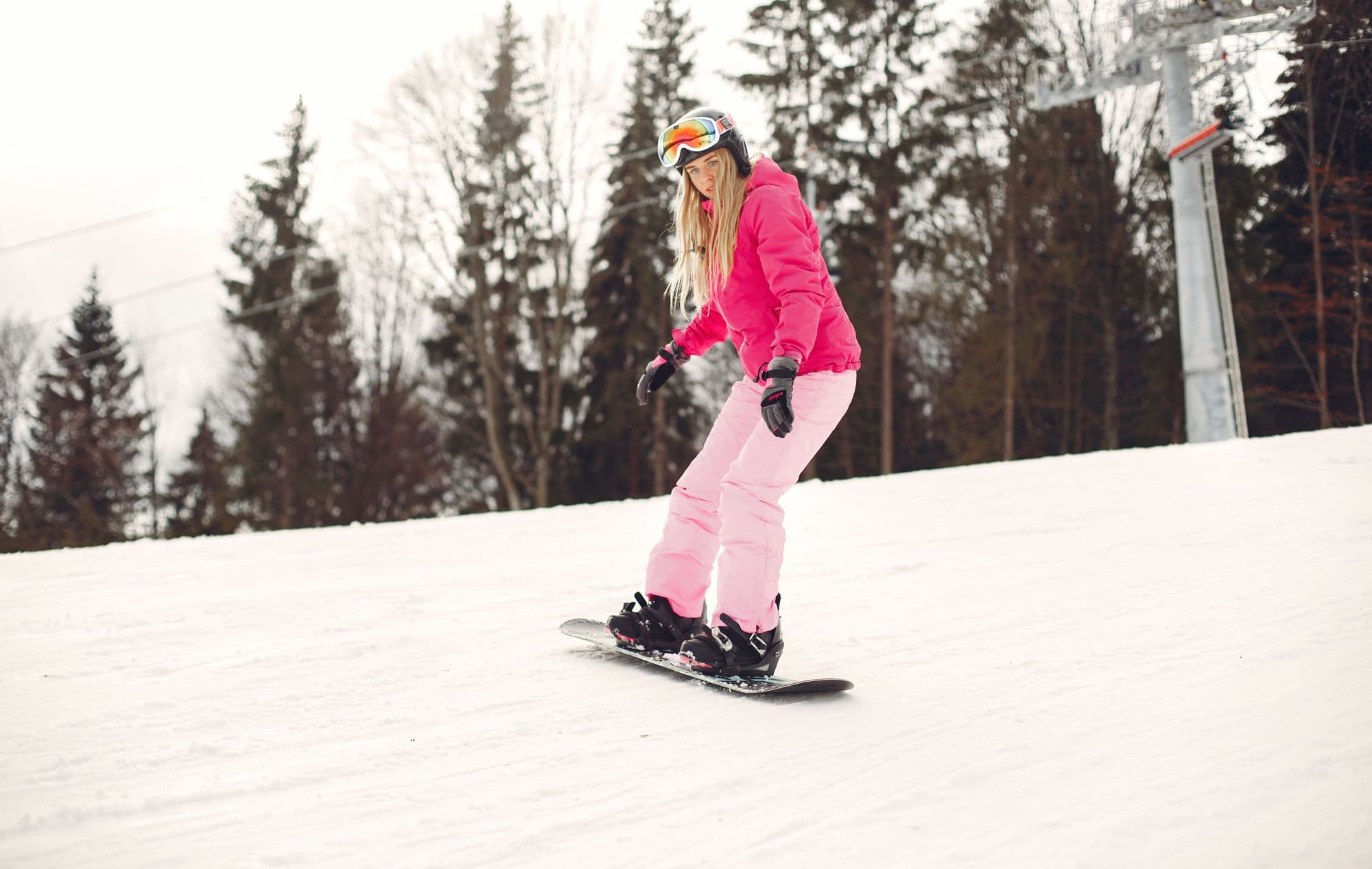 A smiling beginner snowboarder in pink riding down a gentle slope past pine trees