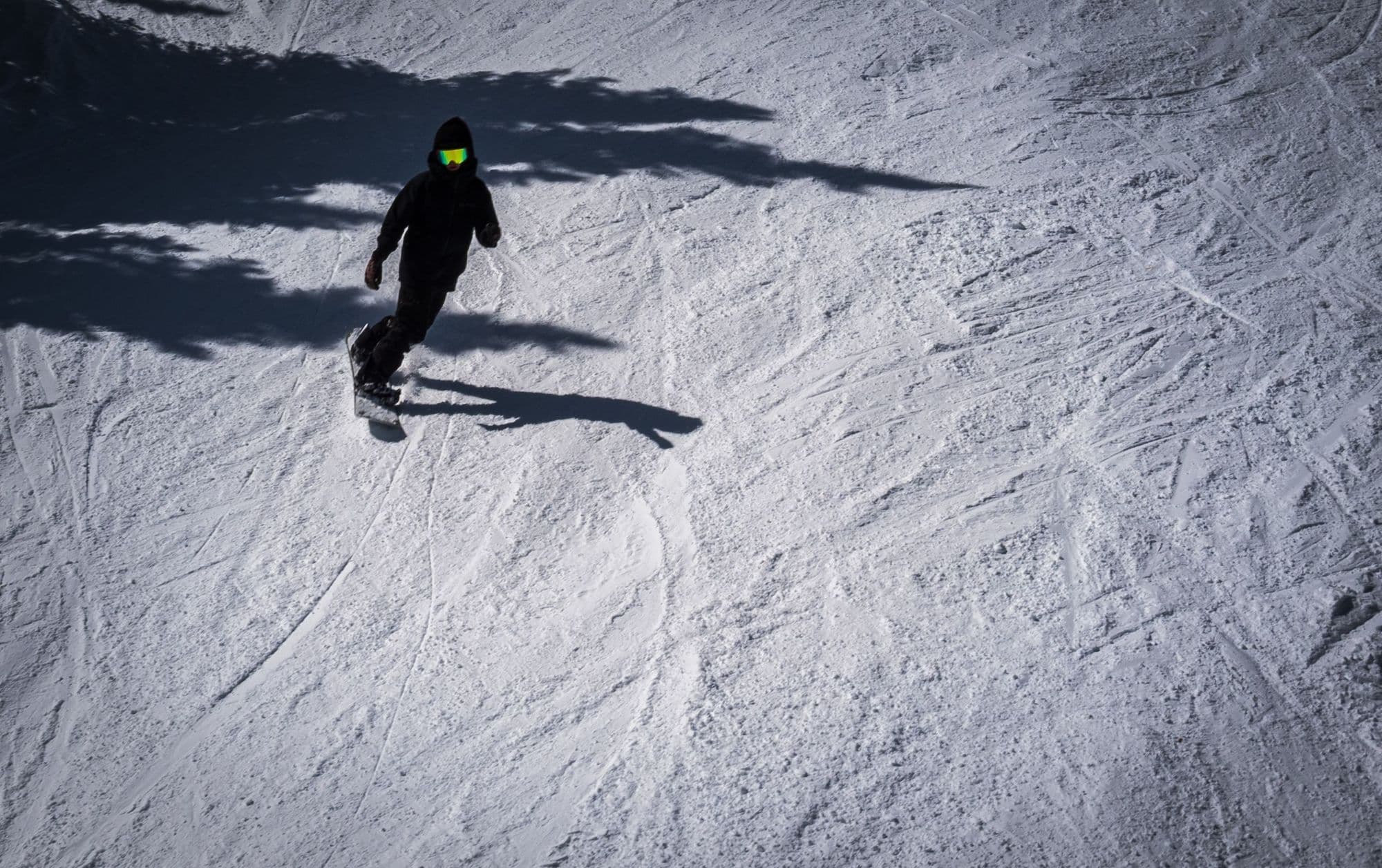 A snowboarder in neon goggles riding down a sunlit slope under tree shadows