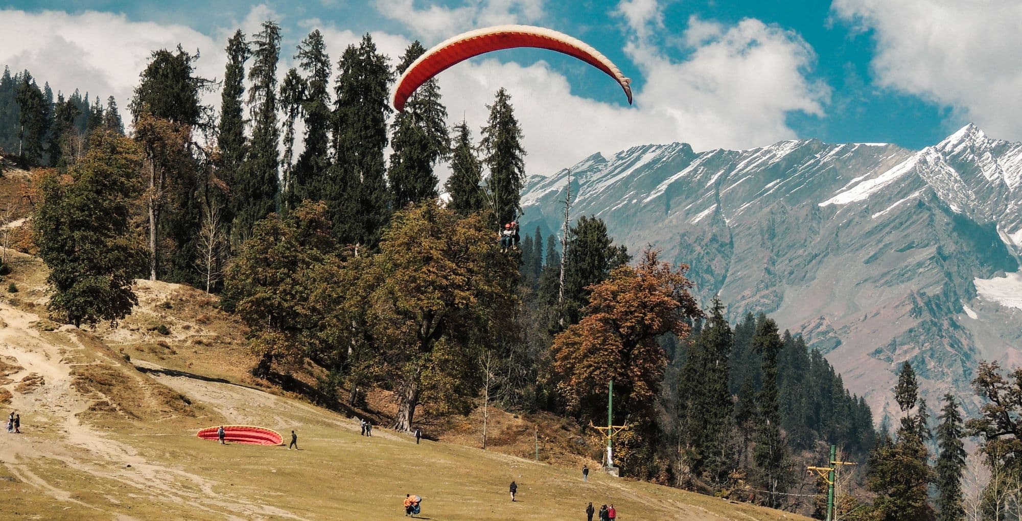 A paraglider launching above pine trees with snowy peaks behind
