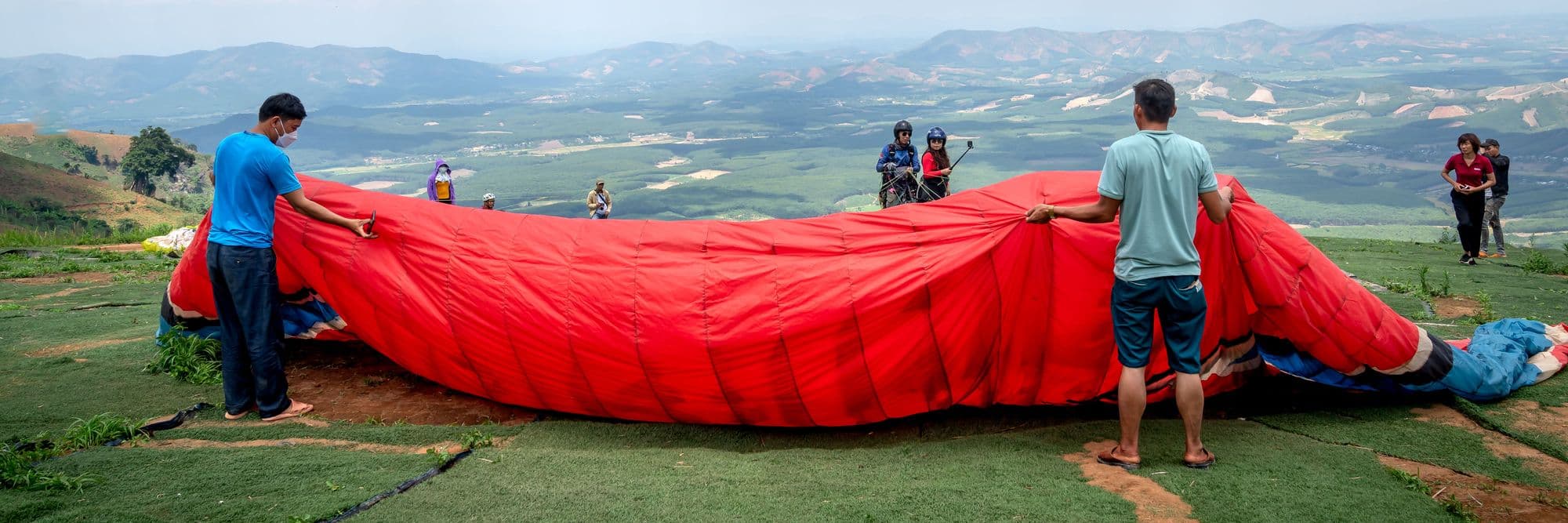 Pilots laying out a red paraglider wing on a hilltop before launch