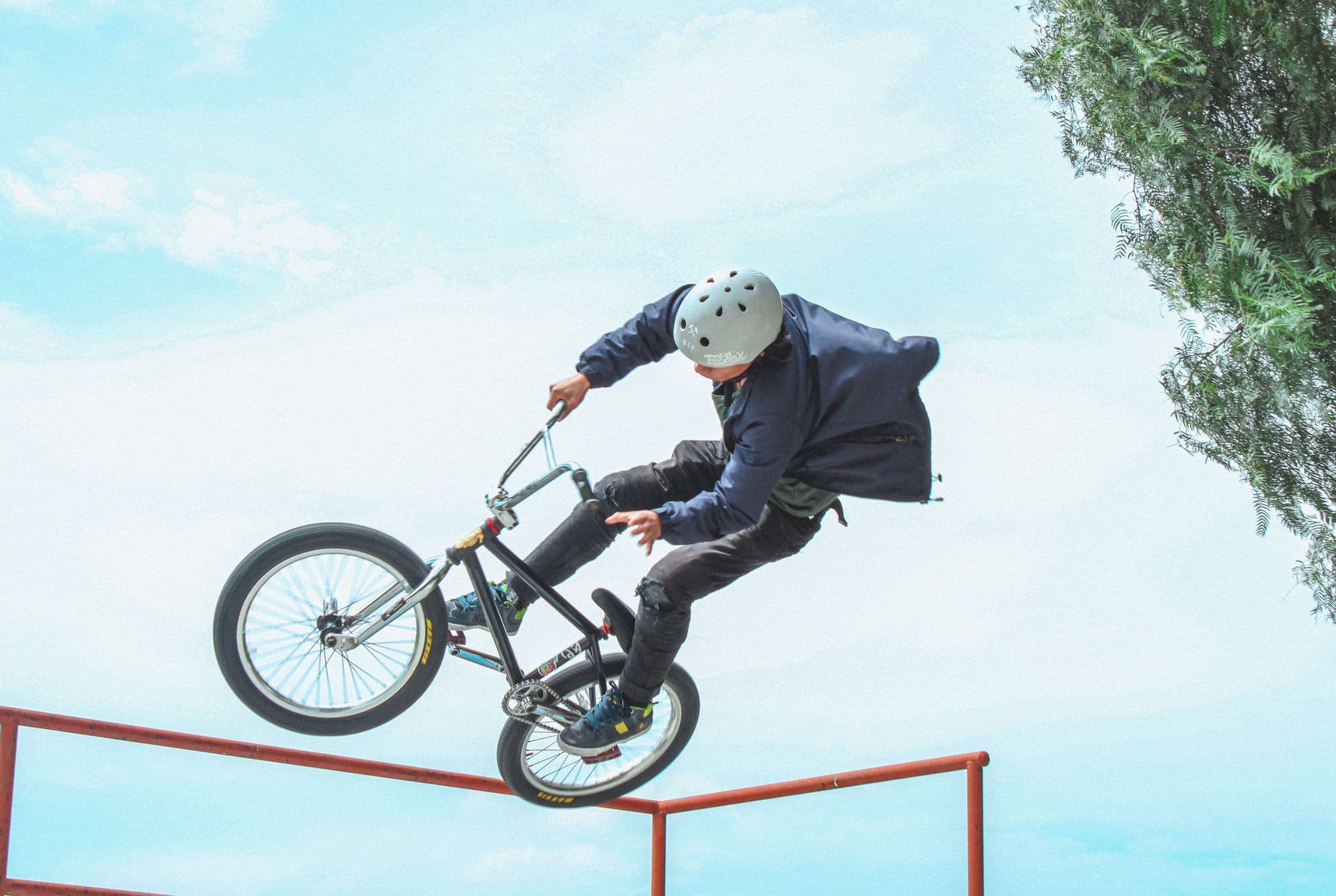BMX rider in a helmet doing a trick over a skatepark rail against a bright sky