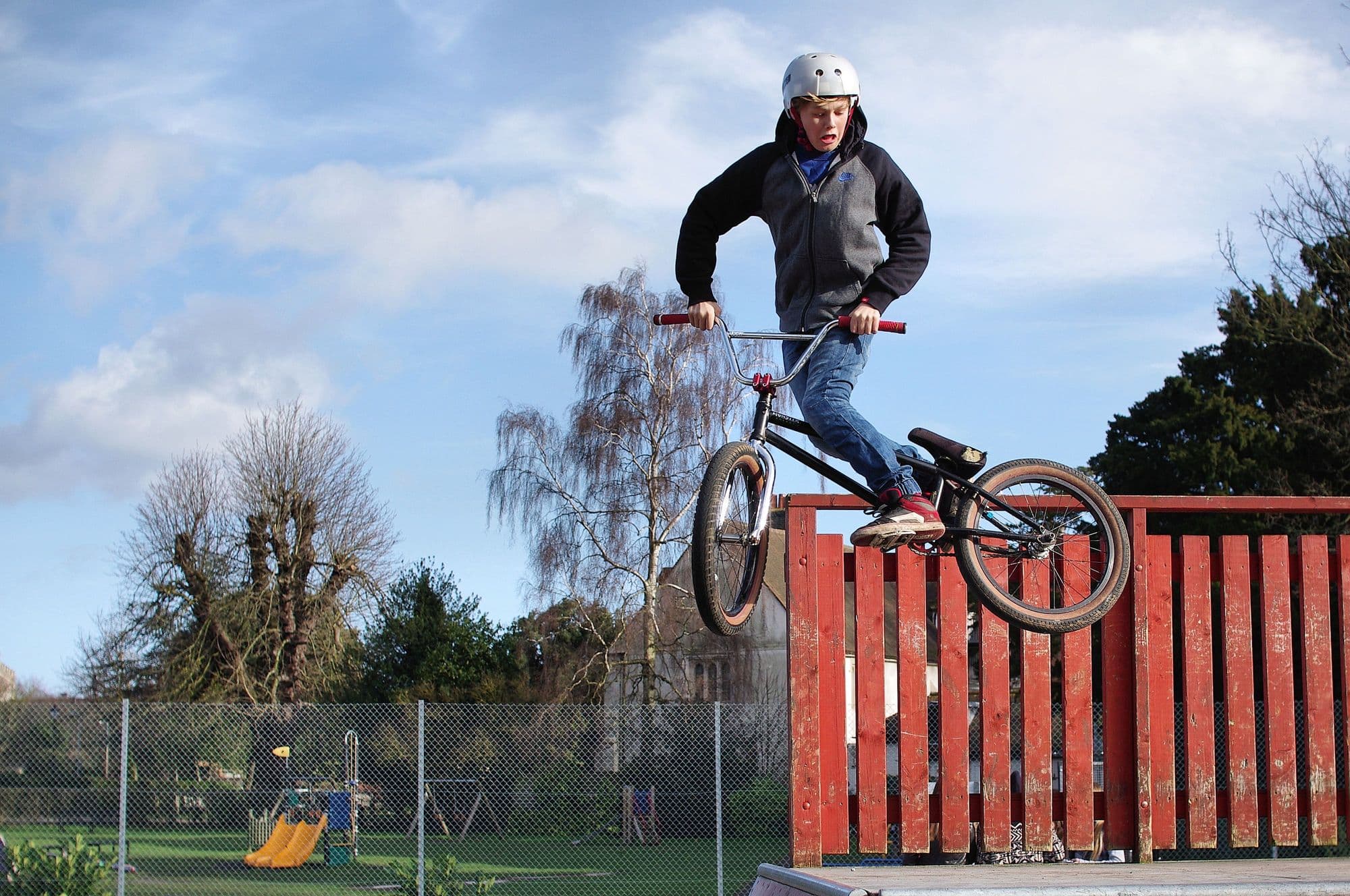 BMX rider in a helmet hopping a red wooden obstacle at a local park