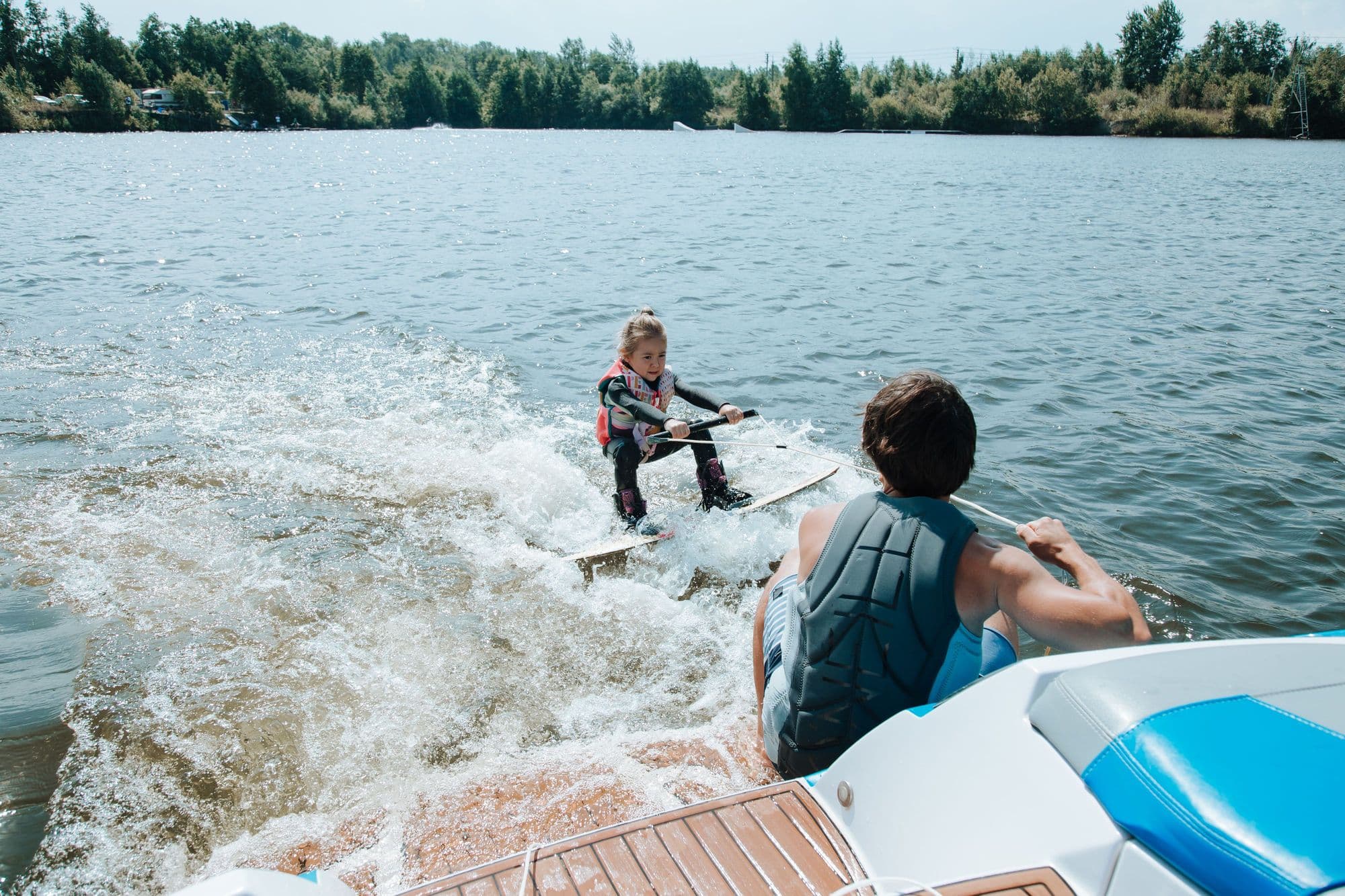 Young rider in a buoyancy vest wakeboarding behind a boat while an adult in a vest watches from the stern