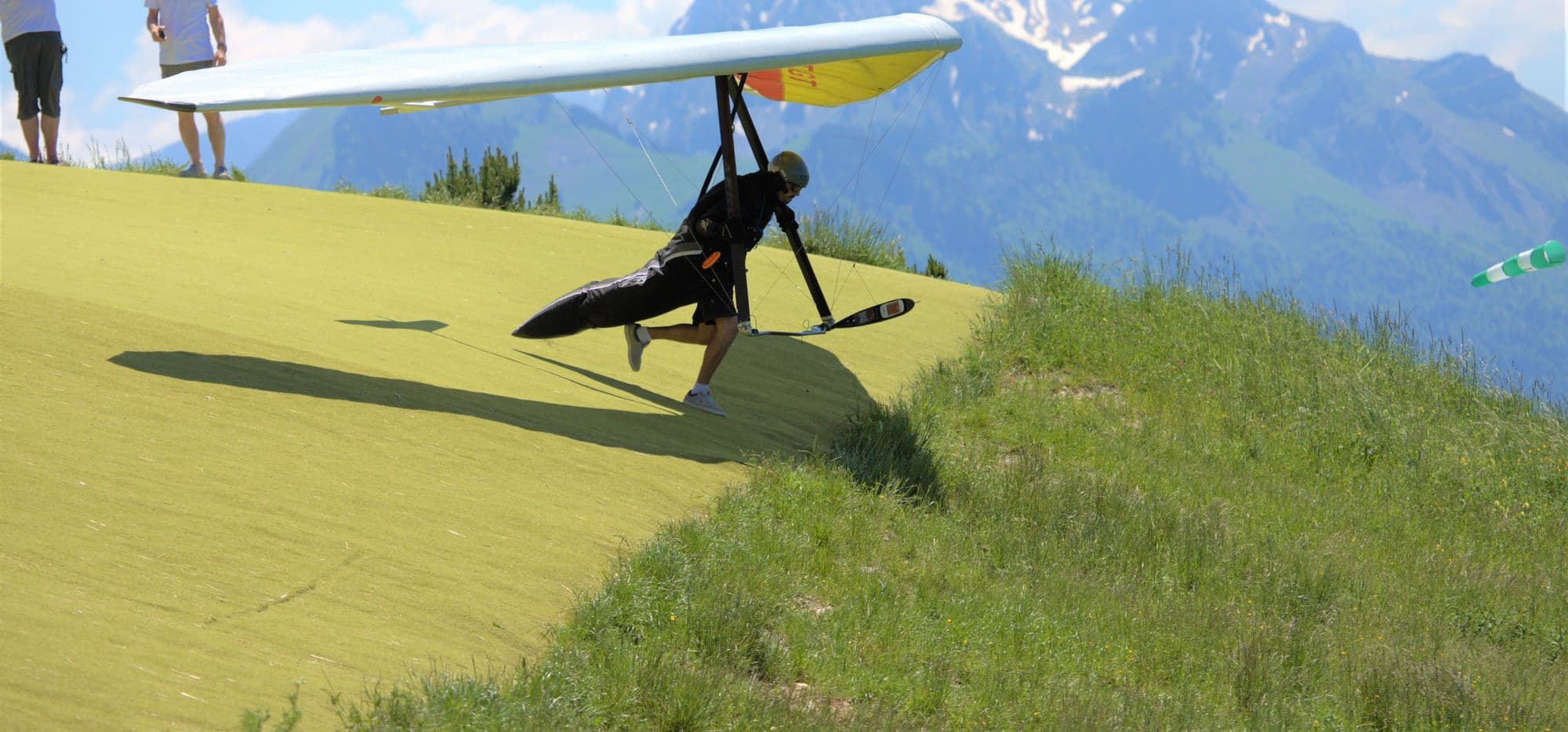 Hang glider pilot running off a grassy hill as onlookers watch