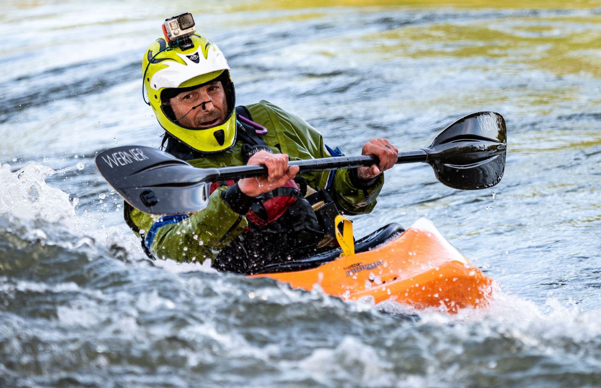 A kayaker in a yellow helmet driving hard through rapids in an orange boat
