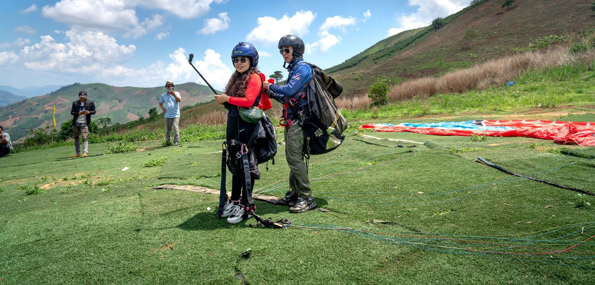 A smiling student pilot in helmet and harness on a grassy launch hill with an instructor