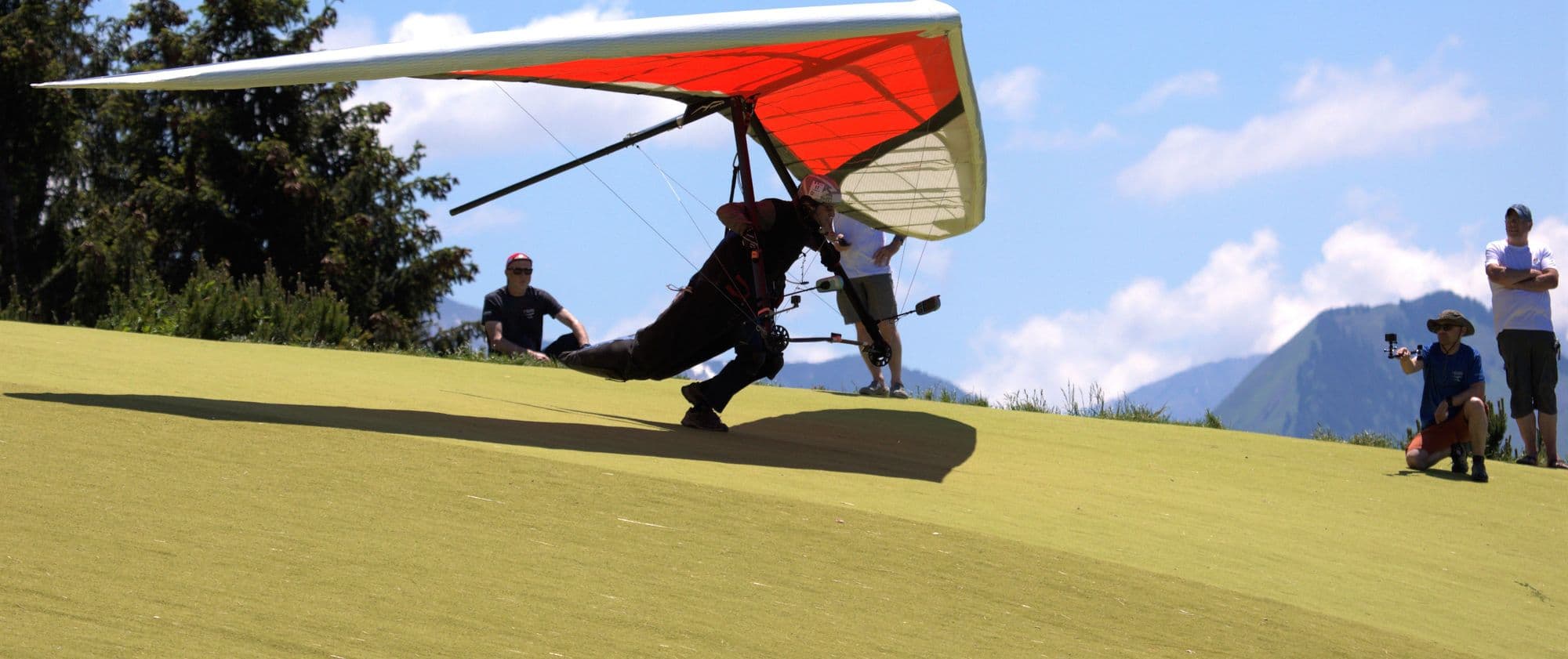 Hang glider pilot launching a red wing while helpers watch from the hill