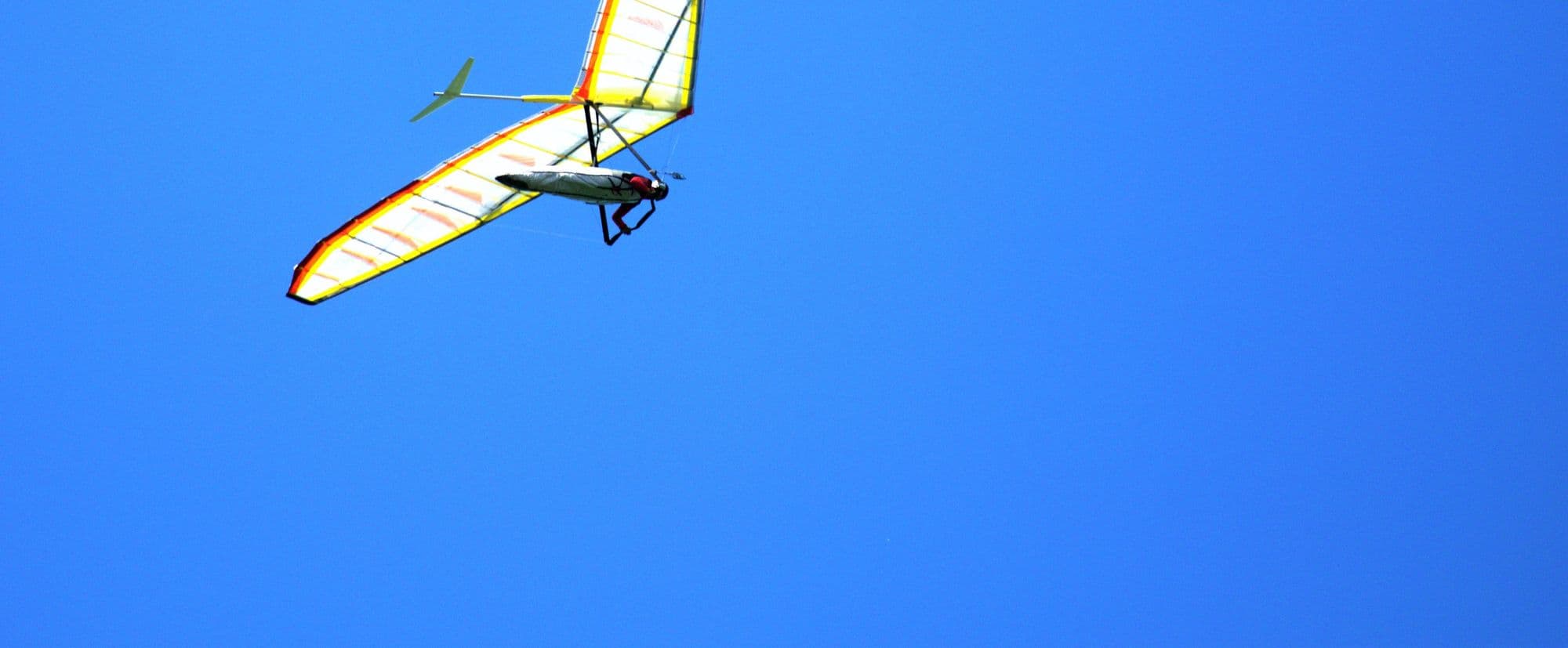 Hang glider with a yellow and white wing flying across a clear blue sky