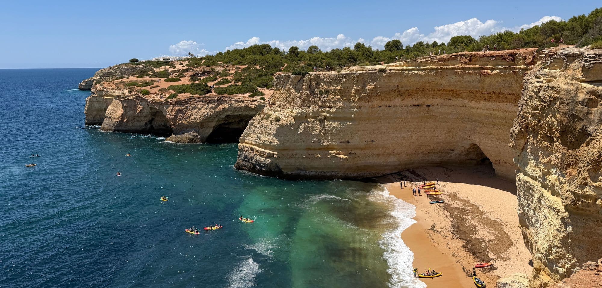 Kayakers paddling past golden cliffs and a sandy cove on clear blue water