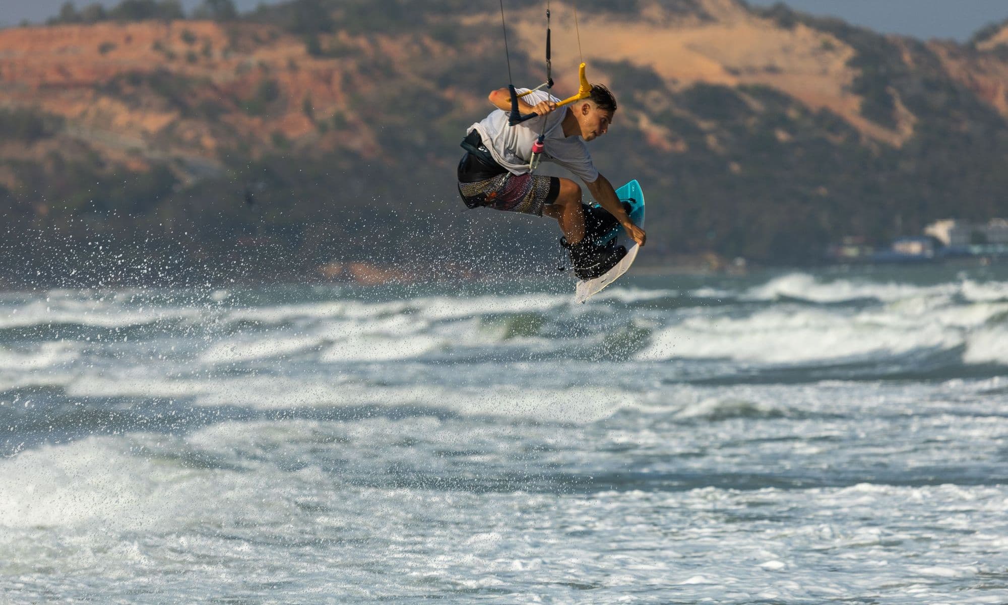 A kitesurfer launching high over breaking waves along a rugged coast