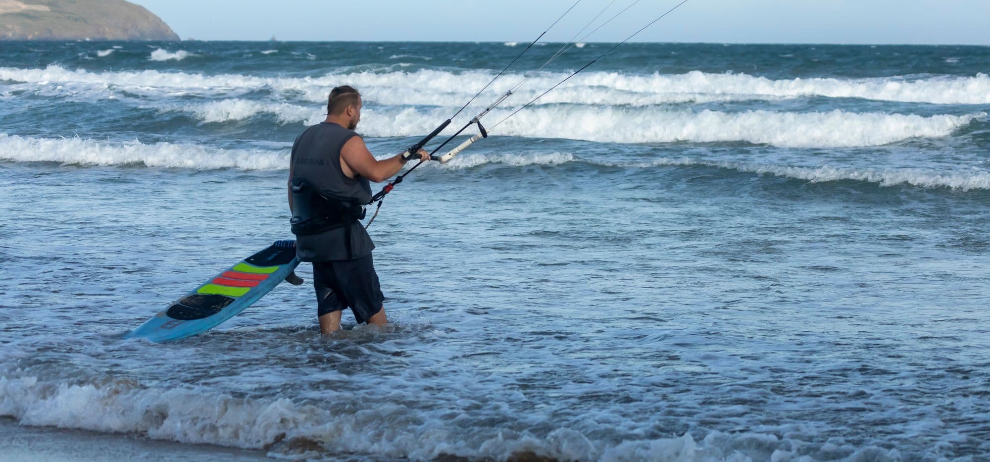 A kitesurfer in a wetsuit walking out through the surf with his board, kite flying