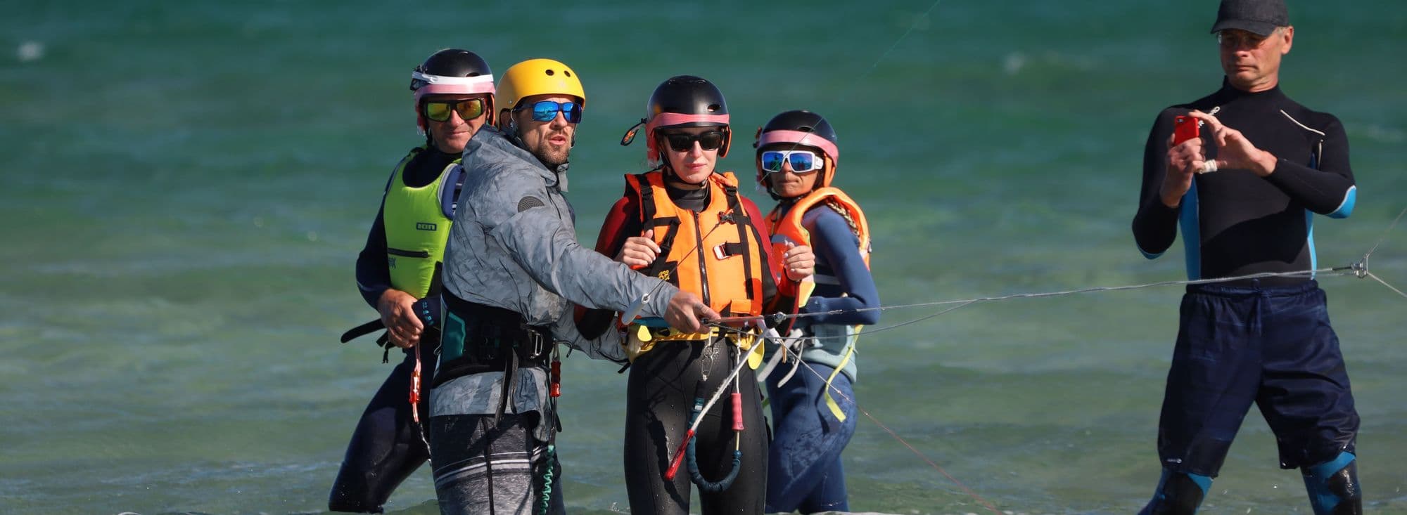 A kitesurf instructor briefing students in helmets and buoyancy aids in shallow water