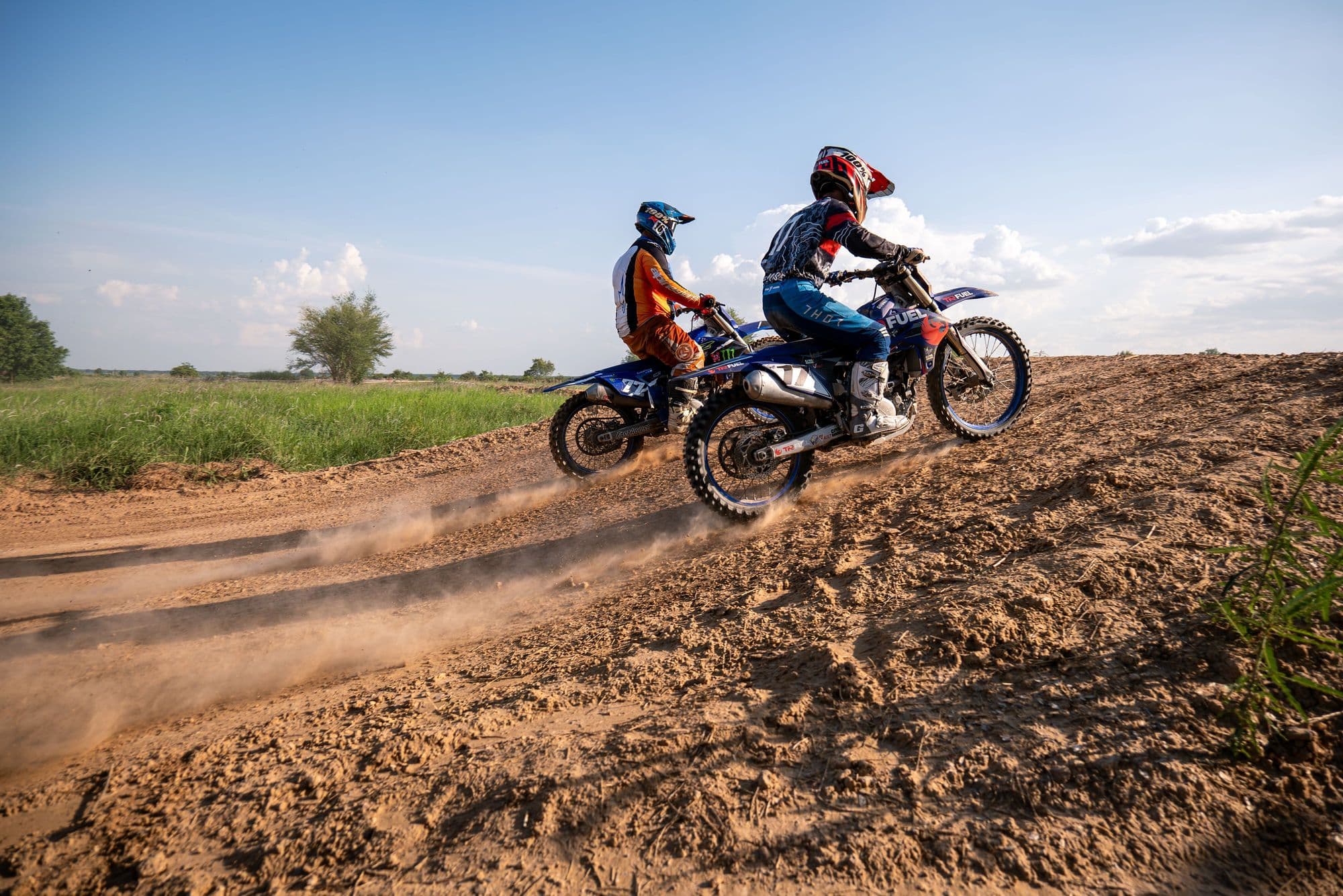 Two motocross riders in full kit racing side by side up a dirt rise under a blue sky