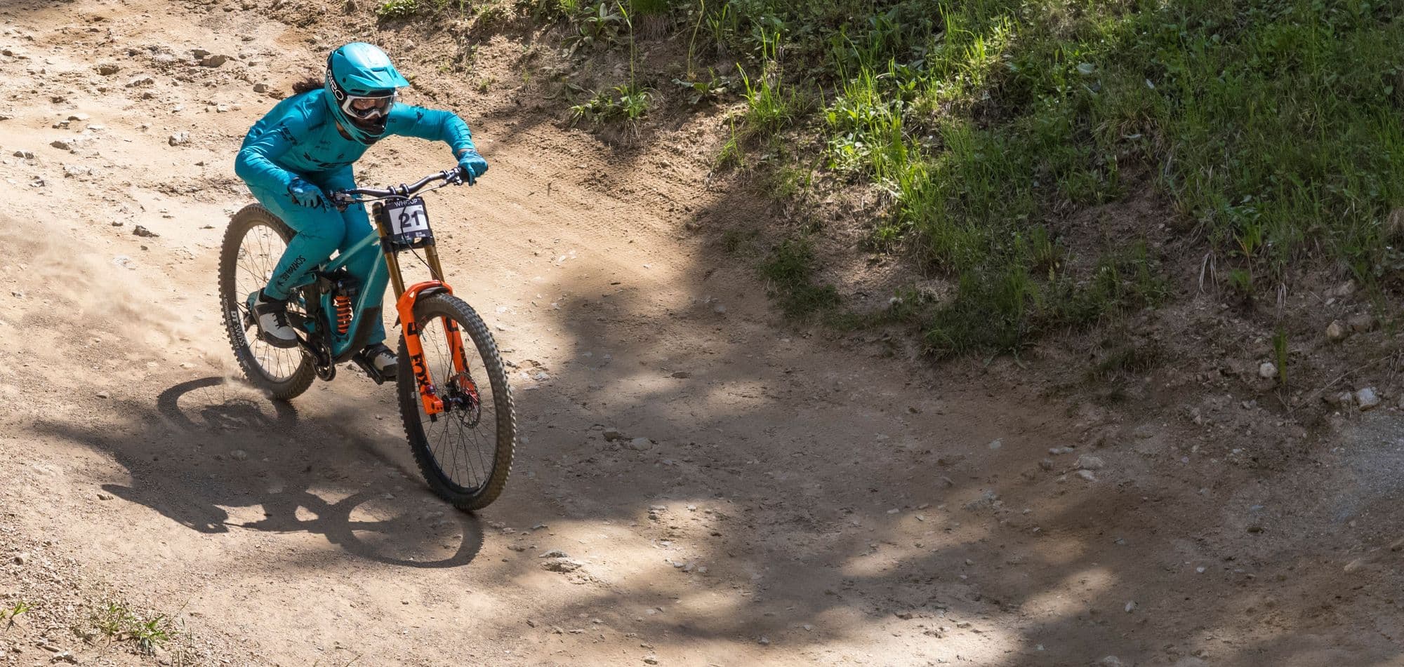 A downhill rider in full-face helmet, goggles and gloves cornering on a sunny dirt trail