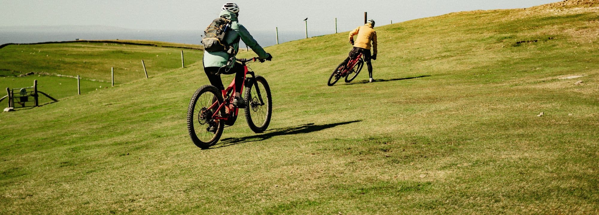 Two mountain bikers in helmets riding across sunny green hills in Wales