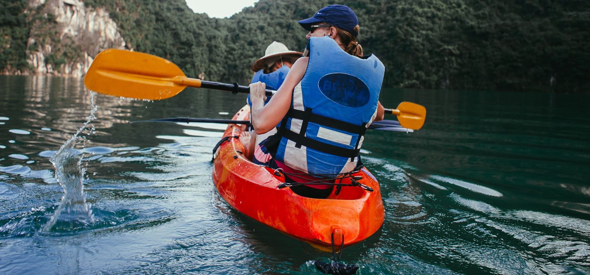Two paddlers in buoyancy aids in an orange tandem kayak below green cliffs