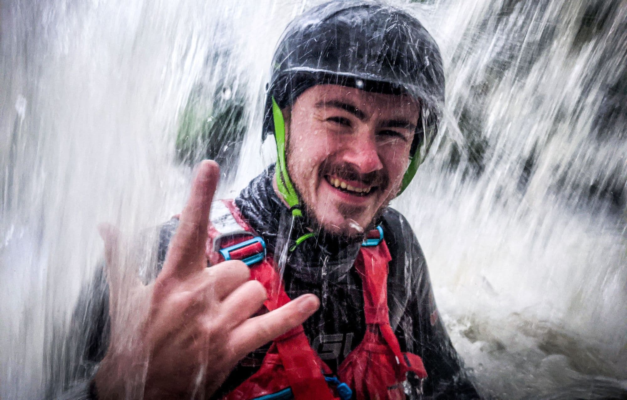 A smiling paddler in helmet and buoyancy aid under a waterfall in Scotland