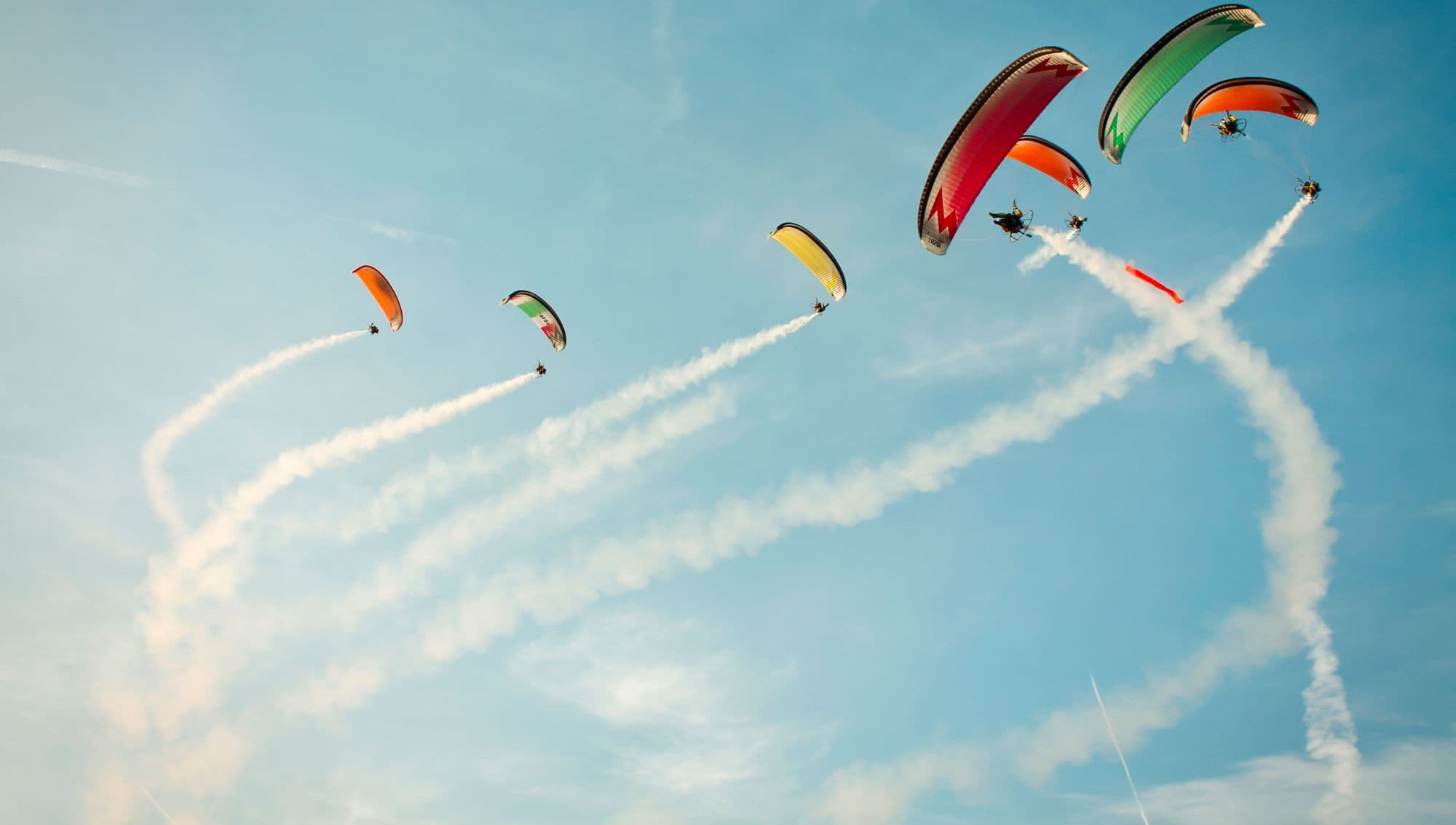 A formation of colourful paragliders trailing smoke across a blue sky