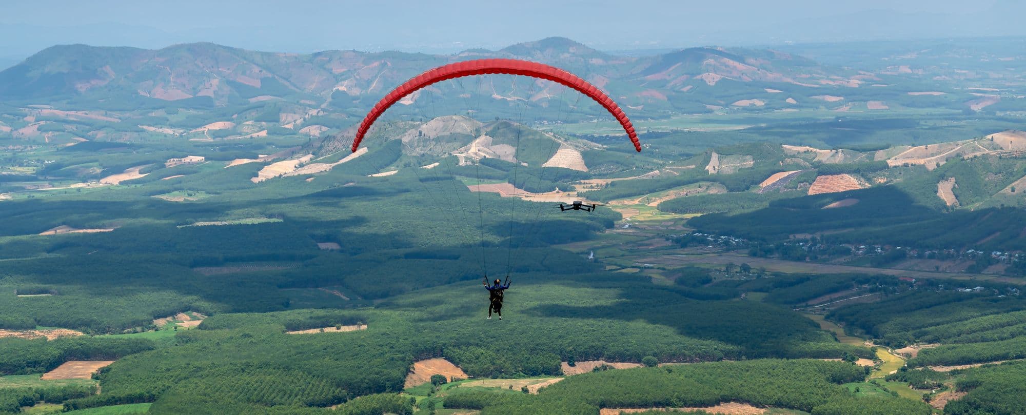 A paraglider under a red wing soaring high above rolling green countryside