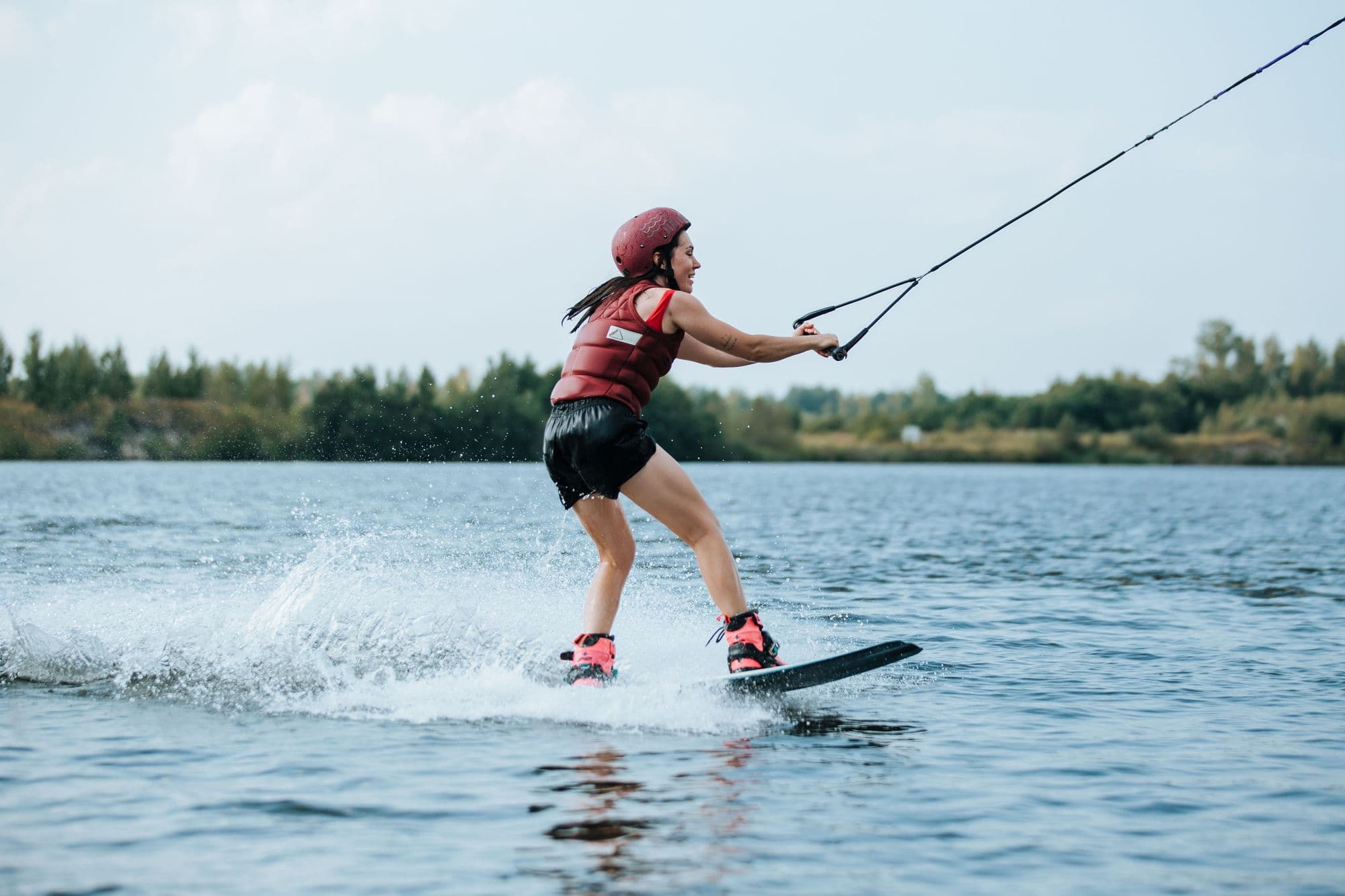 Wakeboarder in helmet and buoyancy vest riding a cable on a tree-lined lake