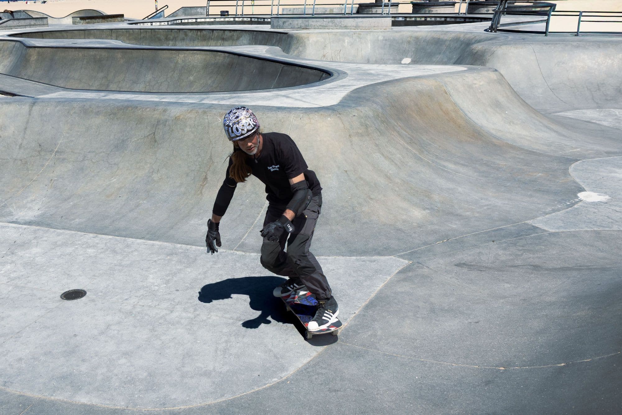 Skater in a helmet, elbow pads and gloves carving through a sunny concrete skatepark