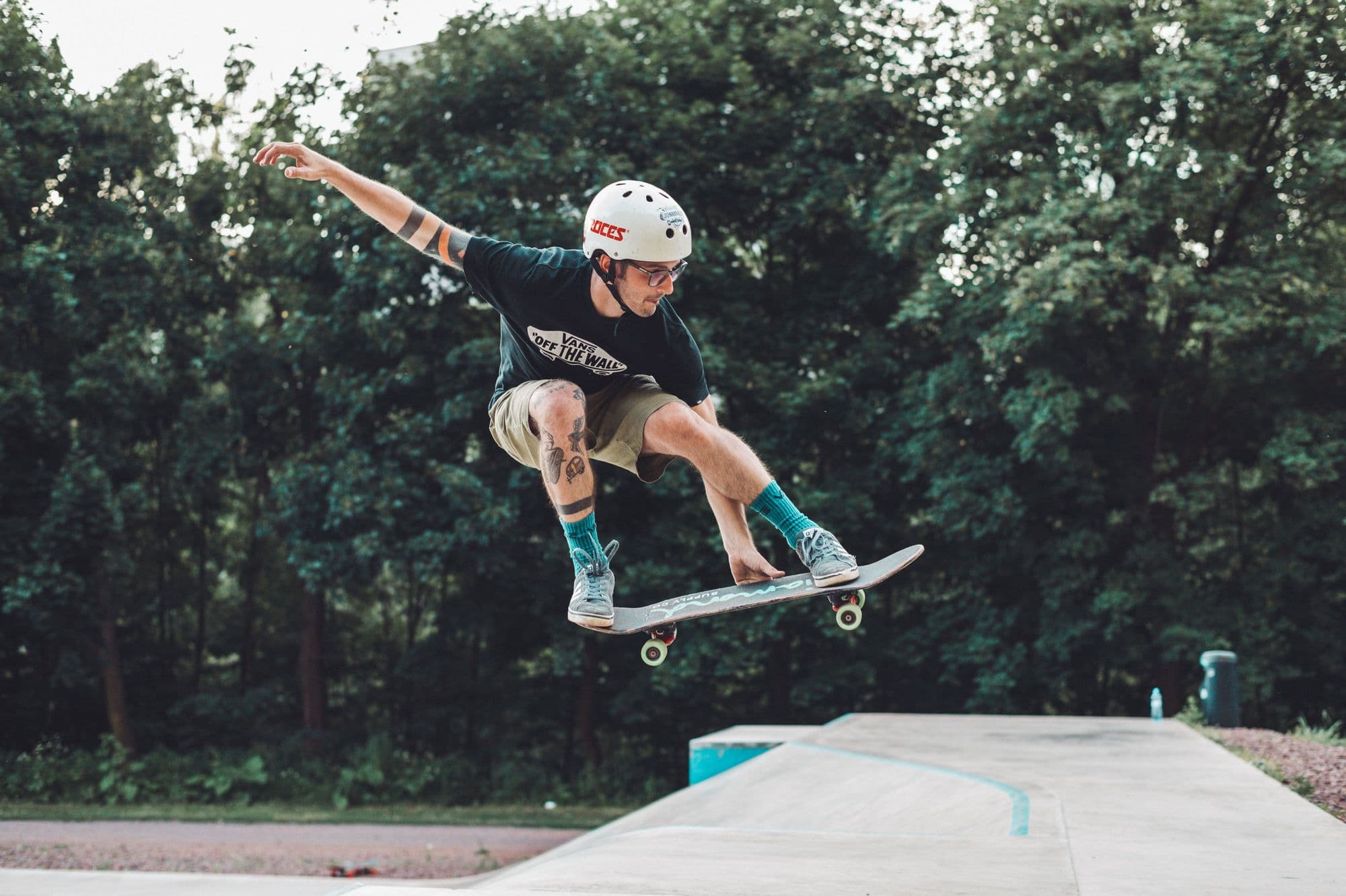 Skater in a helmet flying high off a skatepark ramp against green trees