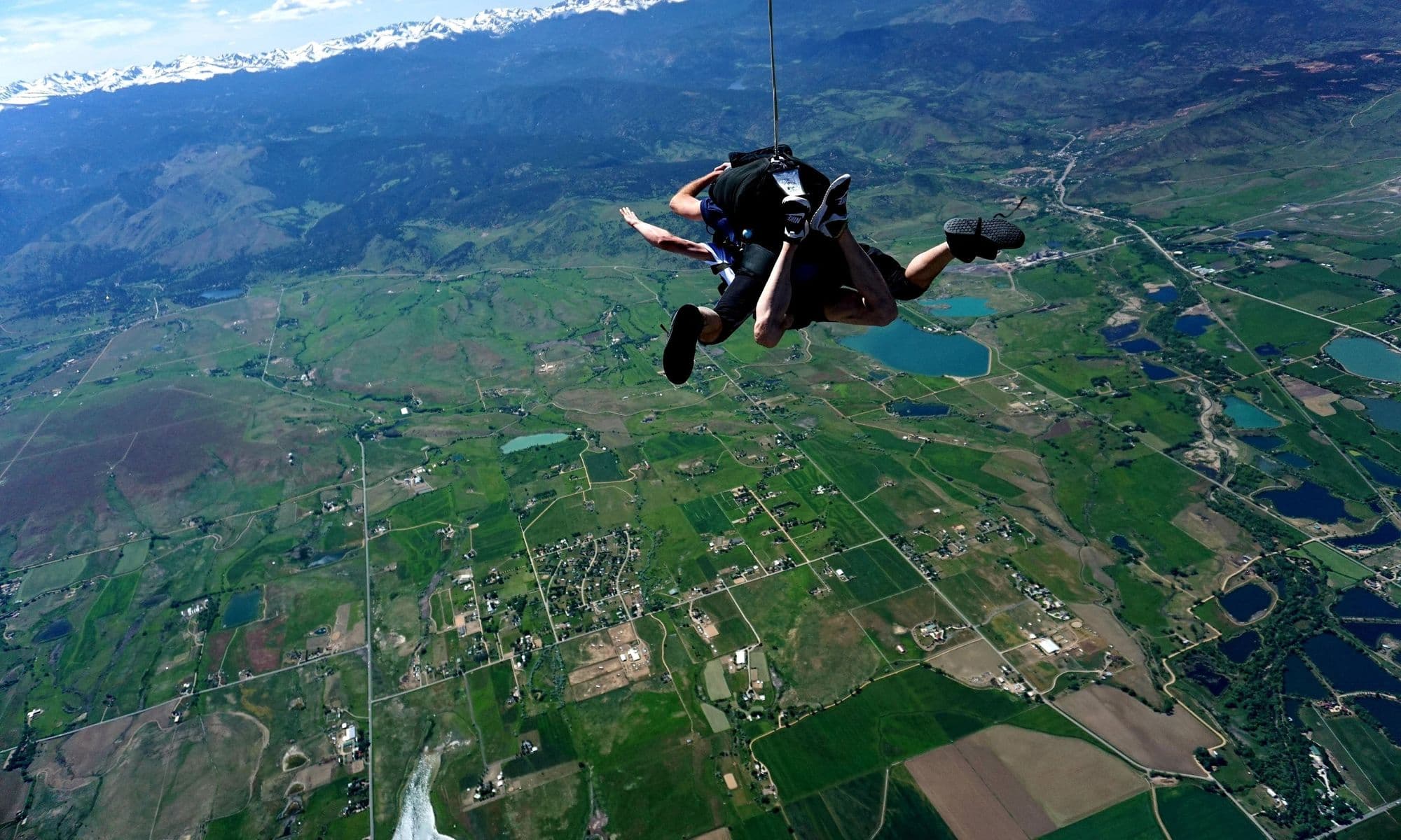 Tandem skydivers in freefall high above green farmland and snowy mountains