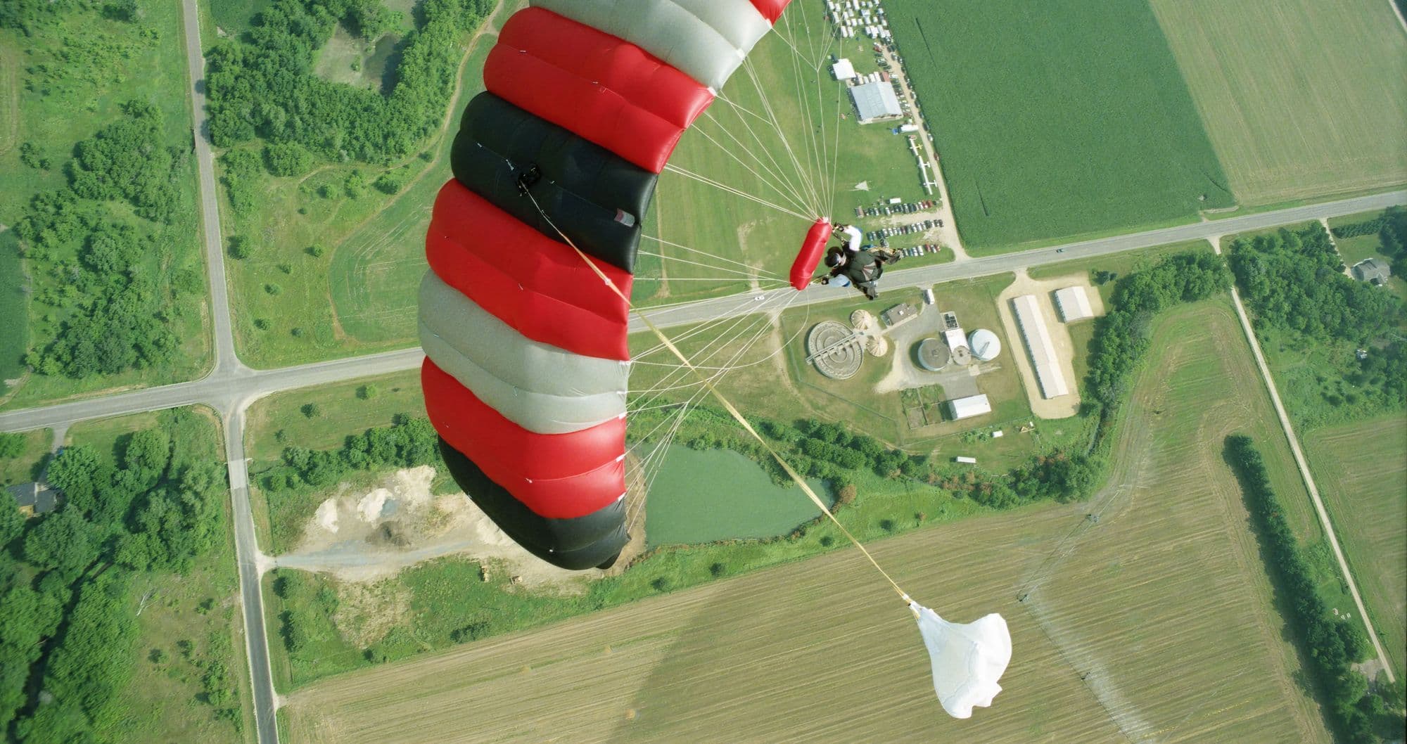 Solo skydiver under a red, white and black canopy above green fields and roads