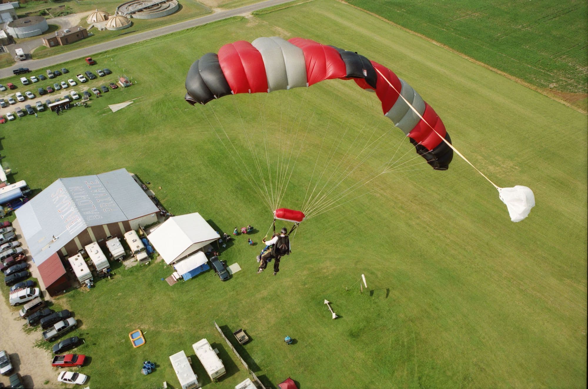 Skydiver under a red and white canopy coming in over a busy drop zone with tents and spectators