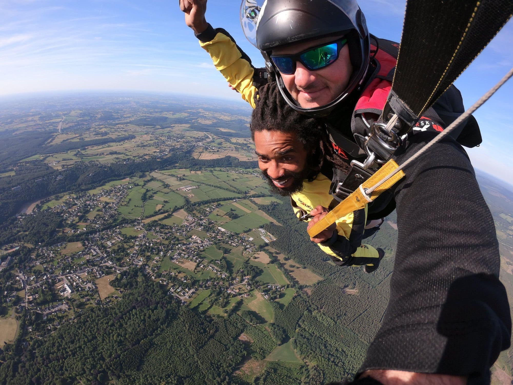 Smiling tandem skydivers in helmets and goggles under canopy over Belgian countryside