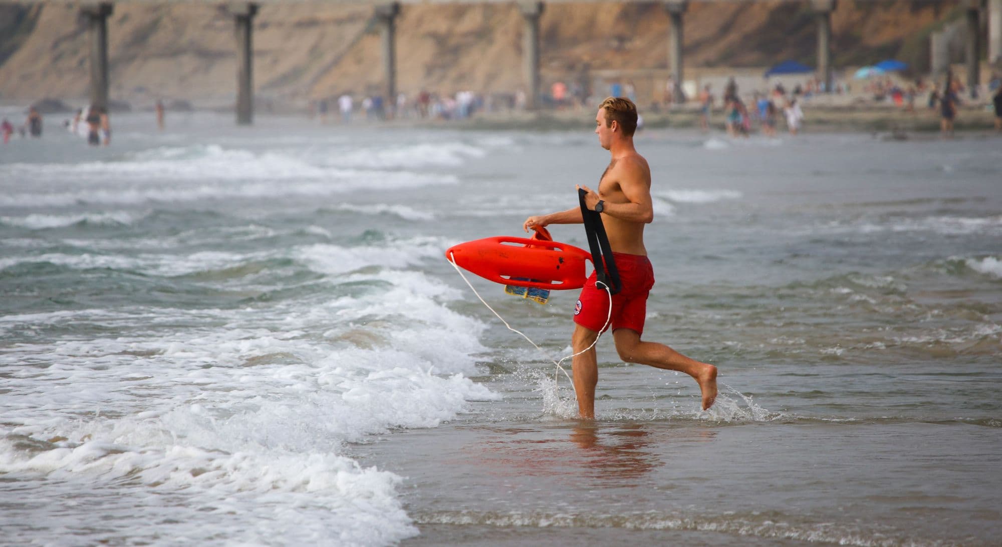 A lifeguard sprinting into the surf with a red rescue tube