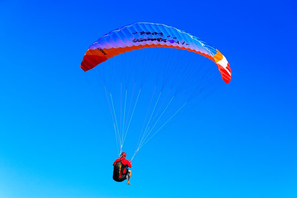 A paraglider in a red jacket and helmet flies under a blue, orange and red wing against a clear blue sky