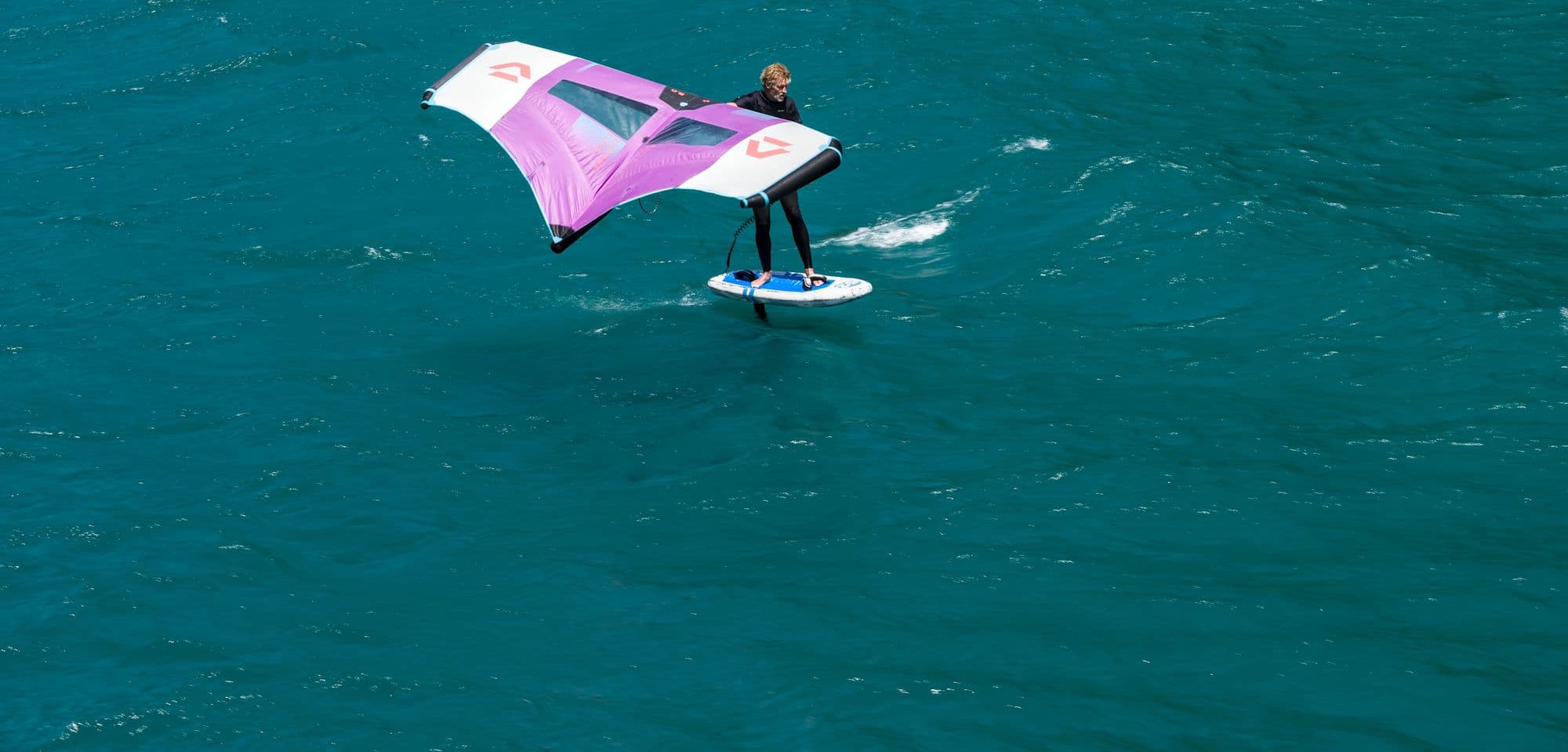 A wing foiler flying above a turquoise lake holding a pink and white wing