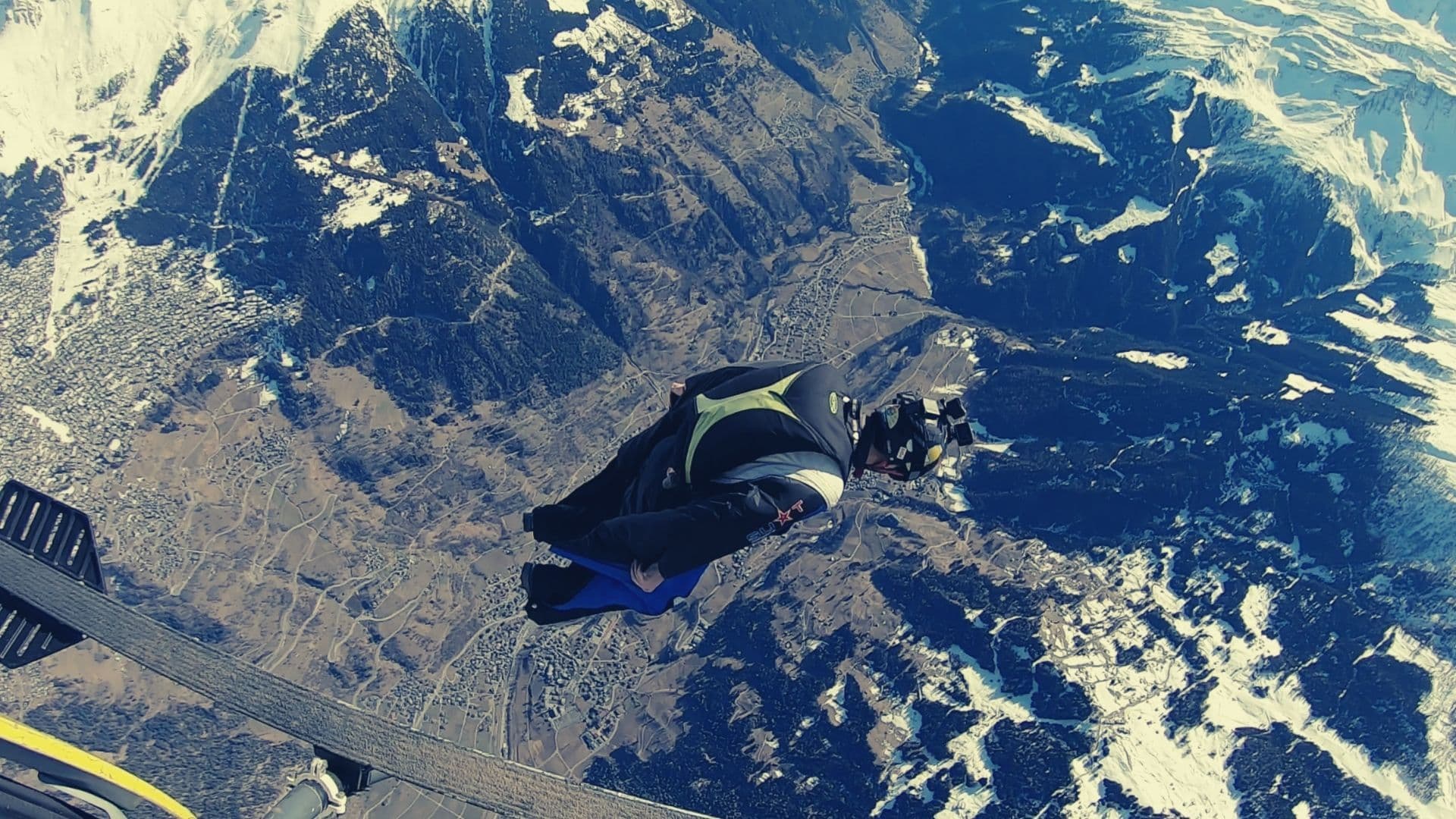 Wingsuit flyer in a helmet gliding away from a helicopter high above snowy mountains