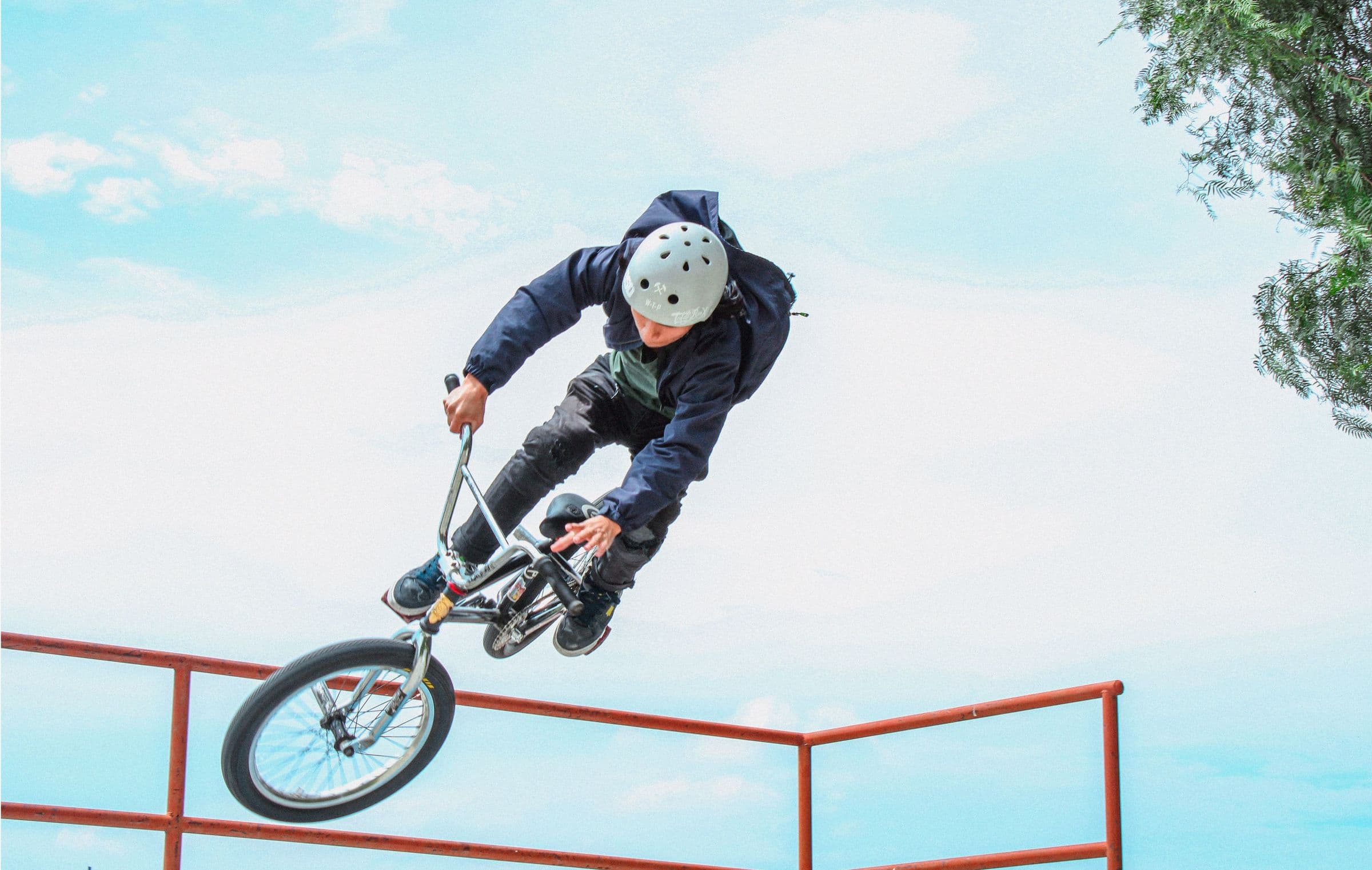 BMX rider in a helmet flying a trick over a skatepark rail under a bright sky