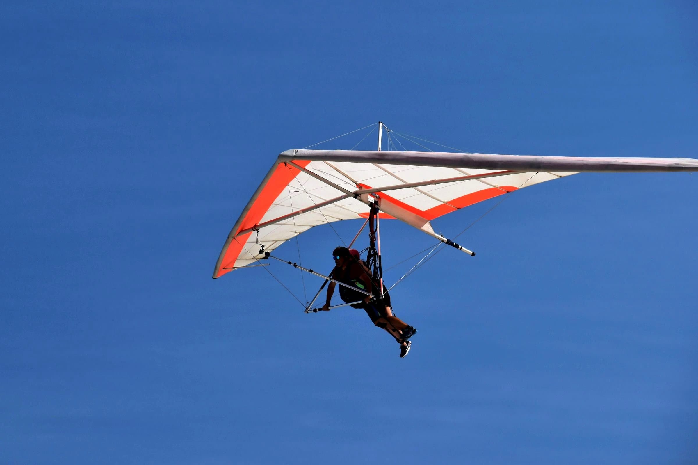A person hangs in a harness under a white wing with an orange-red leading edge, against a clear blue sky