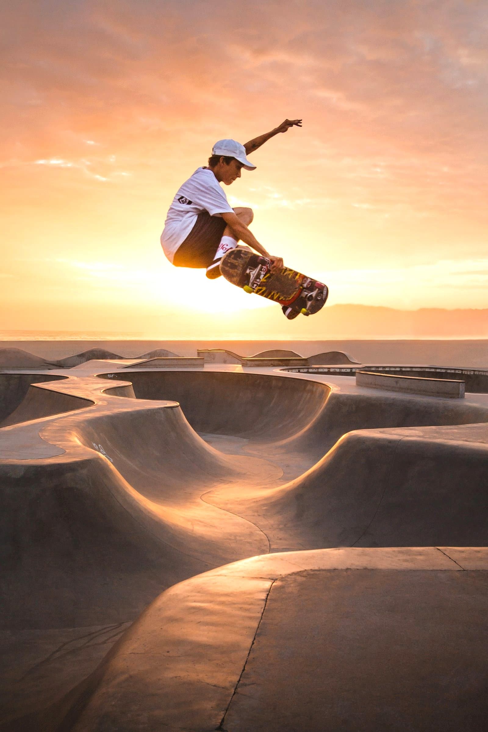 A person in a white cap, white T-shirt and dark shorts grabs a skateboard in the air above a concrete skatepark, with a sunset sky